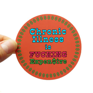 A large round coral sticker with a ring of green dollar signs around the edge. The sticker reads in light blue, magenta pink and green: CHRONIC ILLNESS IS FUCKING EXPENSIVE. The last S is represented with a dollar sign. The sticker is held on the left side with a white thumb & forefinger.
