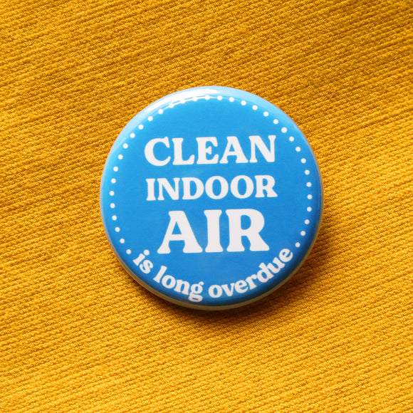 A round medium blue pinback button that reads CLEAN INDOOR AIR, is long overdue in white text. Button is pinned to a yellow fabric.