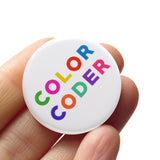 Round pinback button that reads COLOR CODER. Mulit-colored text on a white background. Button is held in a hand