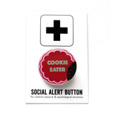 Round pink pinback button with a chocolate  cookie shape that reads COOKIE EATER in mint green text. The button looks like a bite was taken, with crumbs left behind.. Pinback button is on Social Alert Button Backing Card.