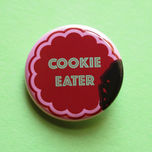 Round pink pinback button with a chocolate  cookie shape that reads COOKIE EATER in mint green text. The button looks like a bite was taken, with crumbs left behind.. Button on a mint green background.