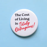 A round white  pinback button that reads THE COST OF LIVING IS TOTALLY OUTRAGEOUS in black & red text,  in two fonts, with a retro 80's vibe.  Button is on a baby blue background.