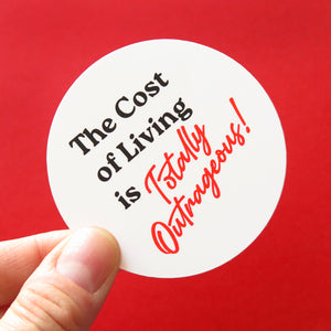 A round white sticker that reads in black text THE COST OF LIVING IS, and TOTALLY OUTRAGEOUS in a red retro 1980's font. Held in the corner by a thumb & forefinger, on a red background.