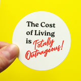 A round white sticker that reads in black text THE COST OF LIVING IS, and TOTALLY OUTRAGEOUS in a red retro 1980's font. Held in the corner by a thumb & forefinger, on a yellow background.