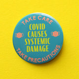 A round scrubs green pinback button that reads COVID CAUSES SYSTEMIC DAMAGE in yellow text, with peach mask icons on either side & a top curve that reds TAKE CARE, an bottom curve that reads TAKE PERCAUTIONS. Button is on a yellow background