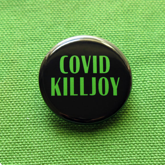 Round black pinback button with green text that reads COVID KILLJOY. Button is on a green woven fabric.