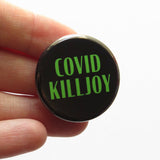 Round black pinback button with green text that reads COVID KILLJOY. Button is held in a white hand.