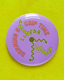 Big 3 inch round lavender pinback button with neon green & coral text laid out like a sick squiggly clock that reads RUNNING ON CRIP TIME, with ON Bisected with A Non Linear Odyssey. Button is on a neon yellow green sweatshirt.