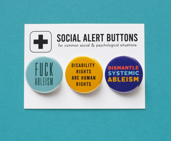 Three round pinback buttons lined up on a SOCIAL ALERT BUTTONS backing card. Reading left to right: A round aqua button reads FUCK ABLEISM, A yellow button that reads DISABILITY RIGHTS ARE HUMAN RIGHTS in black text, and a blue button that reads DISMANTLE SYSTEMIC ABLEISM in red, light blue & yellow.