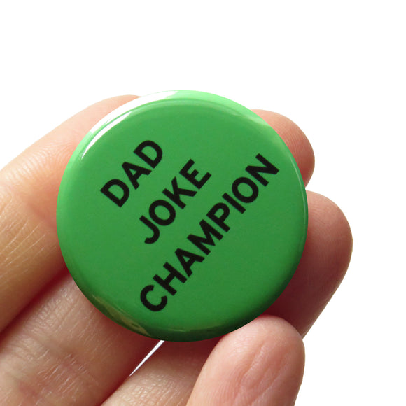 Round pinback button that says DAD JOKE CHAMP. Black text on a green background. Button is held in a hand.