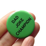 Round pinback button that says DAD JOKE CHAMP. Black text on a green background. Button is held in a hand.