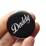 Round shiny black pinback button with a white script font that reads DADDY.  Badge is held in a hand