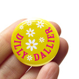 Round, lime green pinback button that reads DILLY DALLIER in deep pink, in a half circle around the edge. The rest of the button has little white daisies with yellow centers. Button is held in a hand.