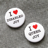 Two round white pinback buttons that read I LOVE QUEER JOY and I LOVE DISABLED JOY in a black serif font, the love being indicated by red hearts. Buttons are on a tiny vintage herringbone patterned fabric.