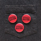 Three round  pinback button that says DISMANTLE SYSTEMIC RACISM. Black text on a neon red background. Buttons are pinned to the pocket of a gray flannel shit.