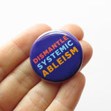 Round, dark blue pinback button that reads DISMANTLE SYSTEMIC ABLEISM in three lines in 3 different colors, red, light blue & yellow. Button is held at the edge of a hand.