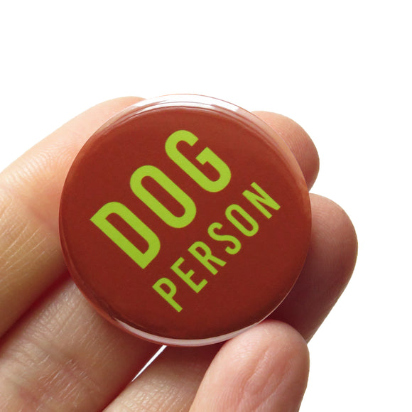 Round reddish-brown pinback button that reads DOG PERSON in lime green san serif text. Button is held in a hand.