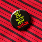 A round black pinback button that reads DONT STOP TALKING ABOUT PALESTINE in green & red text.  Button is on a red & black diagonal stripped shirt
