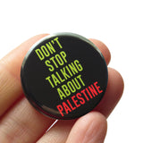 A round black pinback button that reads DONT STOP TALKING ABOUT PALESTINE in green & red text. Button is held in a hand.