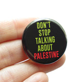 A round black pinback button that reads DONT STOP TALKING ABOUT PALESTINE in green & red text. Button is held in a hand.