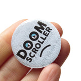 A round pinback button with a pale blue background of faded layered text, with thick black letters that say DOOM SCROLLER, the O's have half lidded eyes, and there's a subtle frown line drown underneath. Button is held in a hand.