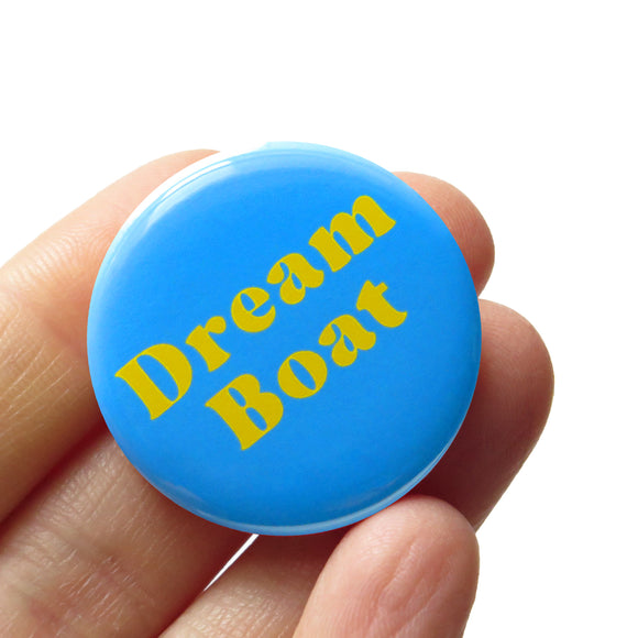 A round light blue pinback button that reads DREAMBOAT in two lines in yellow text. Button is held in a hand.
