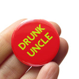 Round red 1.25" pinback button that reads DRUNK UNCLE in light green text. Button is held in a hand.
