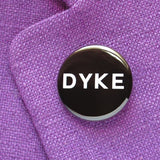 Round, black pinback button that reads DYKE in white text. Badge is pinned to a lavender woven blazer lapel.