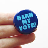 Blue round pinback button that reads EARN MY VOTE! in chunkylight blue text. Button is held on a hand.