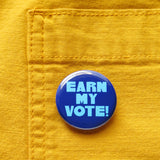 Blue round pinback button that reads EARN MY VOTE! in chunkylight blue text. Button is on yellow pocket.