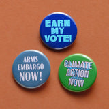 Three round pinback buttons on a rust background: EARN MY VOTE in blues, ARMS EMBARGO NOW in steel gray and white, and Climate Action Now white text on green background.