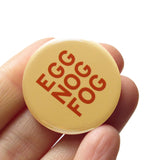 round cream colored 1..25" holiday themed pinback button that reads EGG NOG FOG in nutmegbrown text. Button is held in a hand.