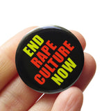 Round black pinback button that reads END RAPE CULTURE NOW in neon yellow and orange san serif text. The button is held in a hand.