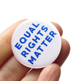 Round white pinback button that reads EQUAL RIGHTS MATTER in blue san serif text. Badge held in a hand.
