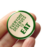 Round cream colored 1.25"  pinback button that reads FEVERYONE DESERVES TO EAT in  green text.  Badge held in a hand.