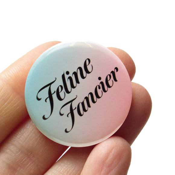 round pinback button that is mint green to peachy-pink ombre with serif black text reading Feline Fancier. Button is held in a hand