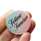 round pinback button that is mint green to peachy-pink ombre with serif black text reading Feline Fancier. Button is held in a hand