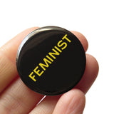 Round black pinback button that reads FEMINIST in yellow san serif text. Edge is in hand.