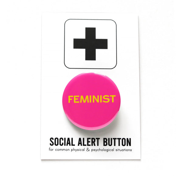 Round pink pinback button that reads FEMINIST in yellow san serif text. Badge is on a Social Alert Button backing card.