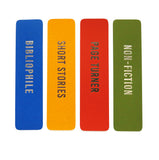 Four richly colored vertical bookmarks with silver or gunmetal embossed text in each, left to right: bright blue that reads BIBLIOPHILE in silver embossed text, marigold yellow bookmark that reads SHORT STORIES in gunmetal embossed foil text, a rich red-organge bookmark that reads PAGE TURNER in gunmetal embossed foil, and a deep moss green bookmark that reads NON-FICITON in gunmetal embossed foil.
