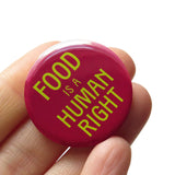 Round burgundy 1.25"  pinback button that reads FOOD IS A HUMAN RIGHT in lime green text.  Badge held in a hand.