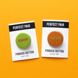 Two pinback buttons on two backing cards with a center perforation that is torn along the perforation line. Button on the left reads FROG with a light green background and brown text. Right sided button reads TOAD in light green with a brown background. Carded buttons are on a golden yellow background.