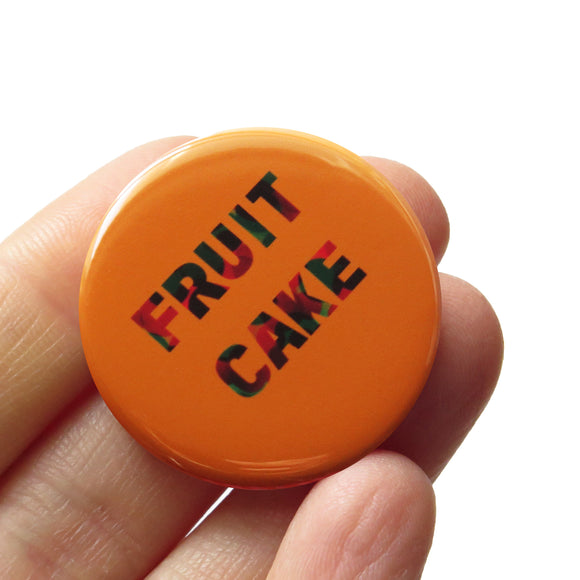 Round caramel colored pinback button that reads FRUIT CAKE in a red green swirl. Button is held in a hand.