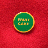 small circular enamel pin, green enamel background with gold text that reads FRUIT CAKE across two lines. Gold colored outline. Pin is on a red sweater.