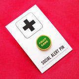 small circular enamel pin, green enamel background with gold text that reads FRUIT CAKE across two lines. Gold colored outline. Pin is on a branded Social Alert Pin backing card.