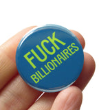 A round teal 1.25" pinback button that reads FUCK BILLIONAIRES. Badge is held in a hand.