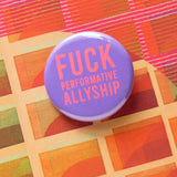 A round medium lavender pinback button with neon red text that reads FUCK Performative Allyship. Button is on an orange patterned surface