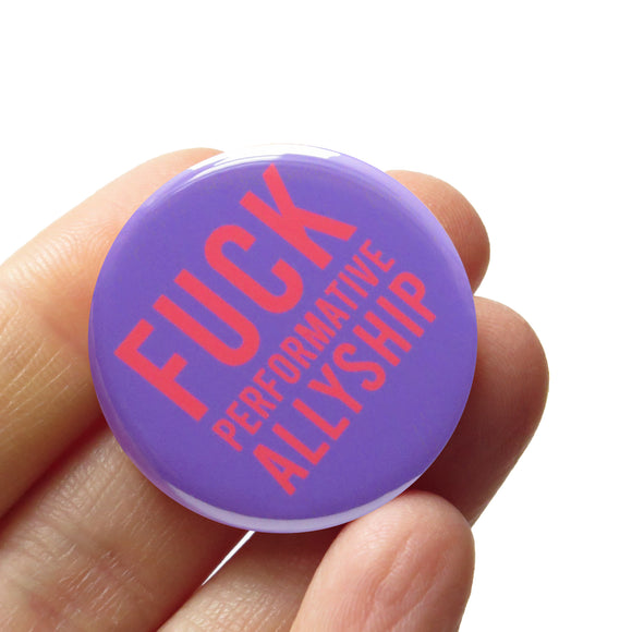 A round medium lavender pinback button with neon red text that reads FUCK Performative Allyship. Button is held in a hand.