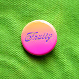 A peach to pink ombre pinback button that reads FRUITY in a purple scripty font with a slight neon yellow drop shadow.  Badge is pinned to a vivid green canvas bag.