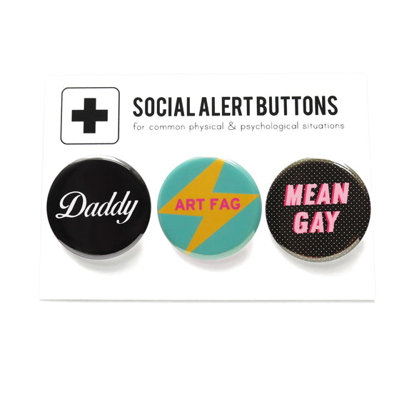 Three pinback buttons lined up on a SOCIAL ALERT BUTTONS backing card. Reading Left to Right: A black pinback button that reads Daddy in a white script font, A teal button with a yellow lightening bolt that reads ART FAG in pink, and a black button with tiny white dots that reads MEAN GAY in light pink with a dark pink drop shadow